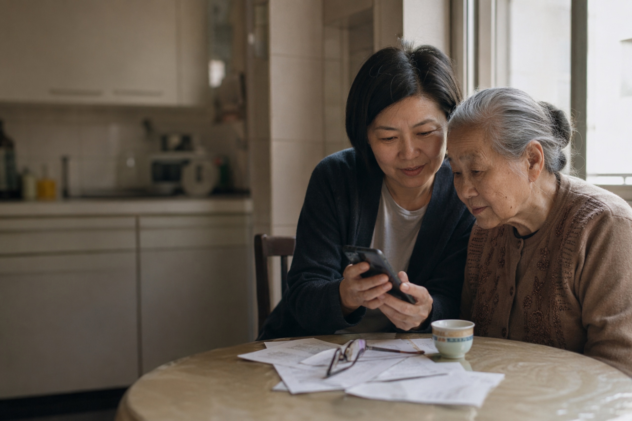 A Chinese adult daughter and her elderly mother looking at a smartphone together at a sunlit kitchen table